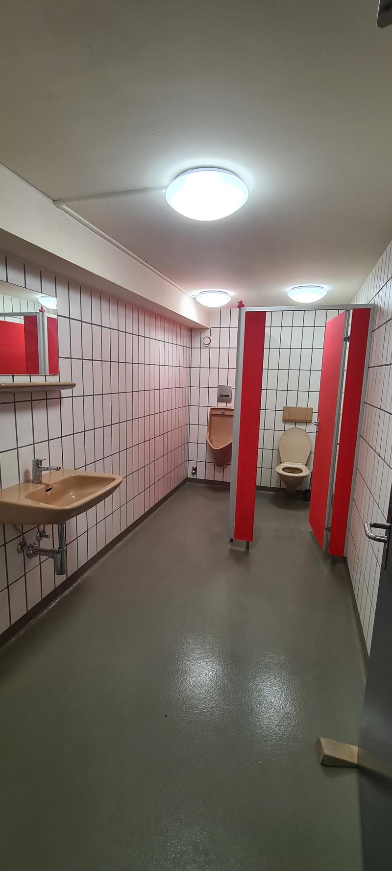 The image shows a public bathroom with white tiled walls, red partitions, a sink, and a toilet. The floor is a gray concrete material. The lighting is provided by ceiling-mounted fixtures.