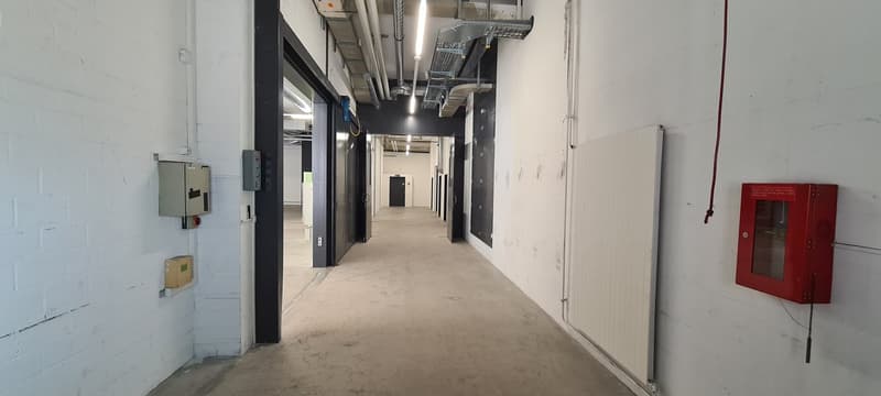 Long, narrow hallway with concrete floor, white walls, and overhead lighting. There are several doors along the hallway and some electrical equipment and a fire extinguisher visible.