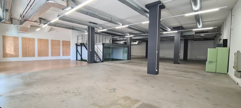 Large open commercial space with concrete flooring, exposed ceiling with ventilation ducts, and wooden panels on the walls. The space appears to be a warehouse or industrial-style office area with a mezzanine level and various equipment and appliances vis