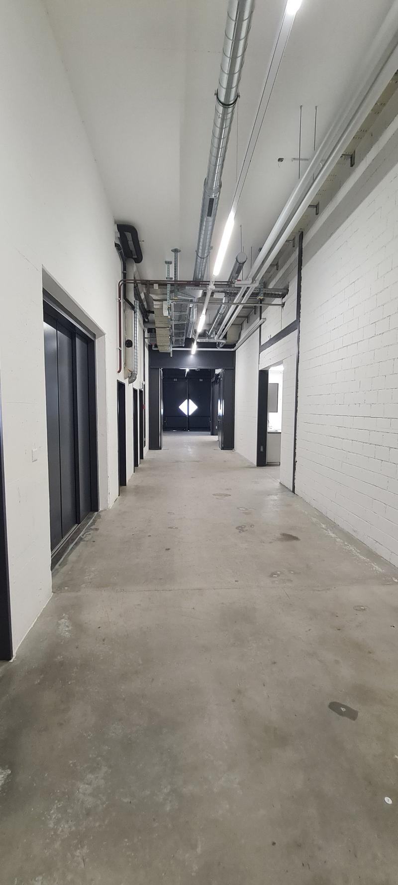 The image shows a long, narrow hallway with concrete floors and white brick walls. The ceiling has exposed ductwork and lighting fixtures. There are several doors lining the hallway, suggesting this is a commercial or industrial space.