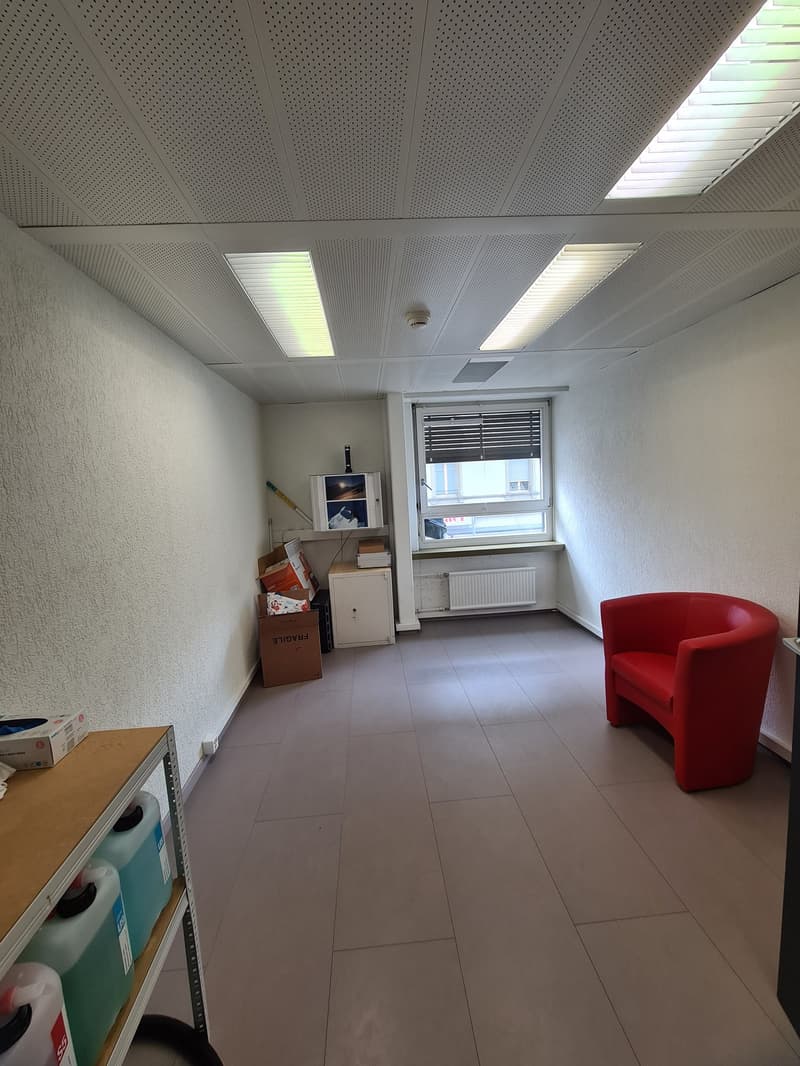 The image shows an office space with a tiled floor, white walls, and a suspended ceiling with recessed lighting. There is a red armchair and some boxes and storage units in the room.