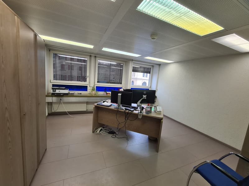 The image shows an office space with a desk, computer monitors, and other office equipment. The room has a tiled floor, gray walls, and a suspended ceiling with lighting fixtures. The windows have blinds installed.