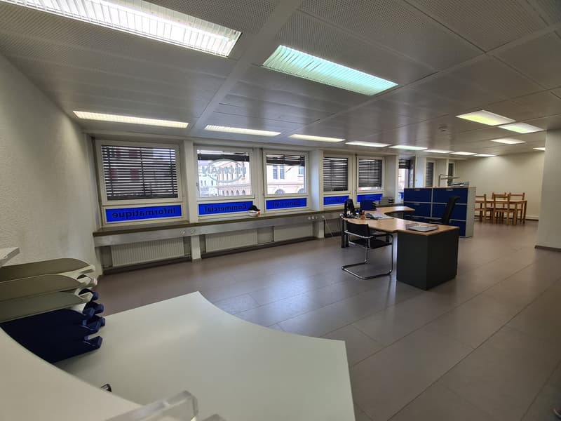 The image shows a modern office space with a reception desk, chairs, and desks. The room has large windows, fluorescent lighting, and a tiled floor. There are blue and white branding elements visible on the walls and furniture.