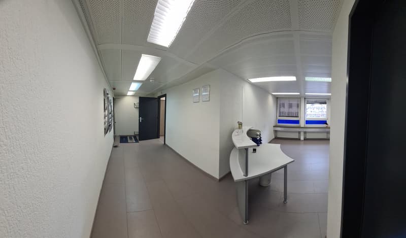 The image shows an office space with a modern and minimalist design. The room has a white ceiling with recessed lighting, gray walls, and a tiled floor. There is a desk with a computer monitor and other office equipment, as well as a door leading to anoth