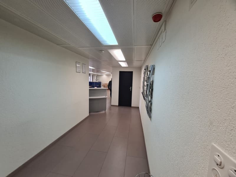 The image shows a hallway or corridor in an office or commercial space. The walls are painted in a light color, and the ceiling has a grid of recessed lighting panels. The floor is covered with light-colored tiles. There is a black door visible at the end