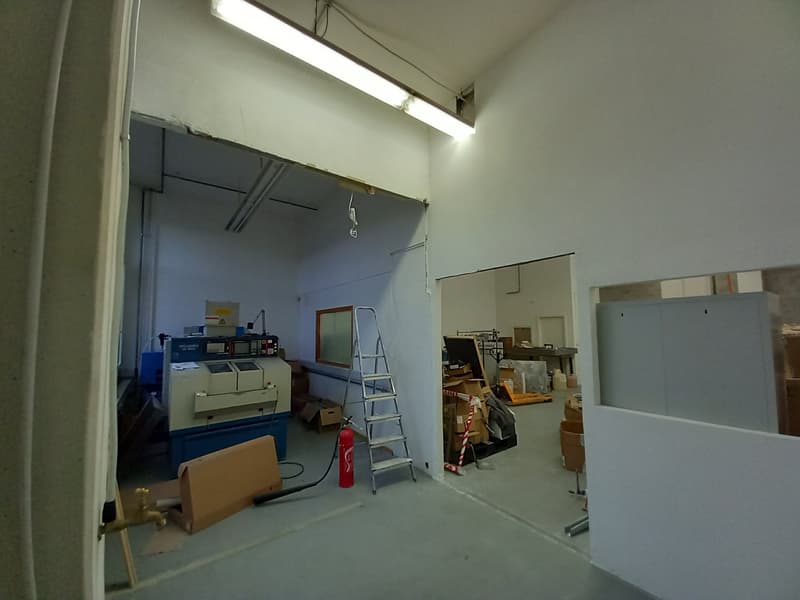The image shows a storage room or warehouse-like space. It contains various office equipment and supplies, including a printer, boxes, and a ladder. The room has white walls and a fluorescent light fixture overhead.