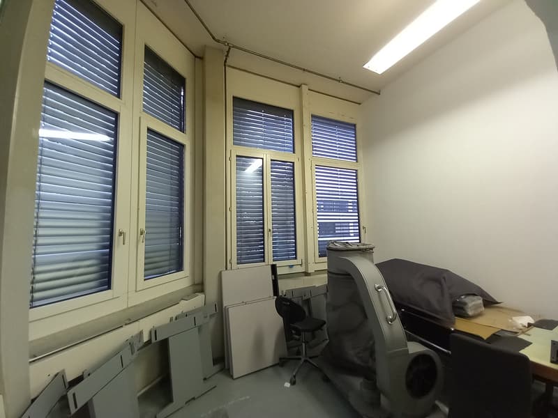 The image shows an office space with large windows, blinds, and office equipment such as a desk, chair, and other items. The room appears to be well-lit and has a neutral color scheme.