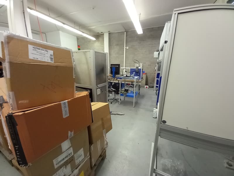 The image shows a storage room or warehouse-like space. There are several cardboard boxes and packages stacked on the floor, and some shelving units or storage cabinets visible. The room has concrete walls and a utilitarian, industrial appearance.