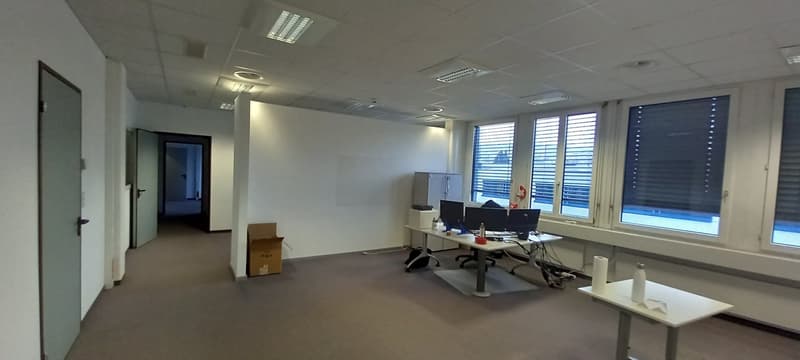 The image shows an open office space with a desk, computer, and other office equipment. The room has large windows, a tiled ceiling, and appears to be a commercial or professional work environment.