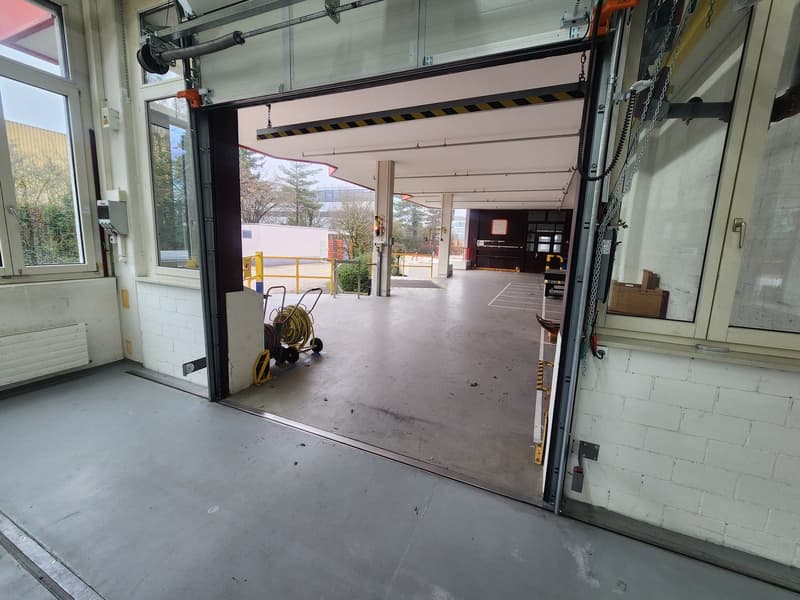 An open garage with grey floor, windows, and white brick walls, with a yellow gate and a trolley and a hose laid out on the floor.