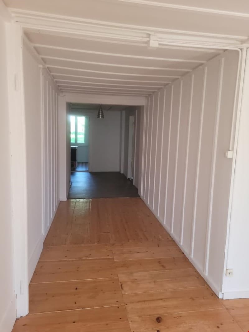 The image shows a long, narrow hallway with white walls and a wooden floor. The hallway appears to be part of a larger space, with a doorway visible at the end leading to another room.