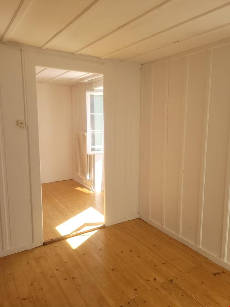 This image shows an empty room with white walls, a wooden ceiling, and a wooden floor. The room has a window that is letting in natural light, creating a bright and airy atmosphere. The room appears to be a simple, minimalist space with no visible furnitu