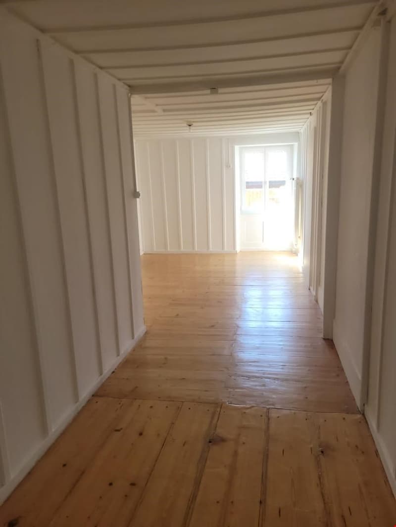 The image shows a long, narrow hallway with white walls and a wooden floor. The hallway has a window at the end, allowing natural light to enter. The ceiling is also white and has a series of beams running across it, creating a clean and minimalist aesthe