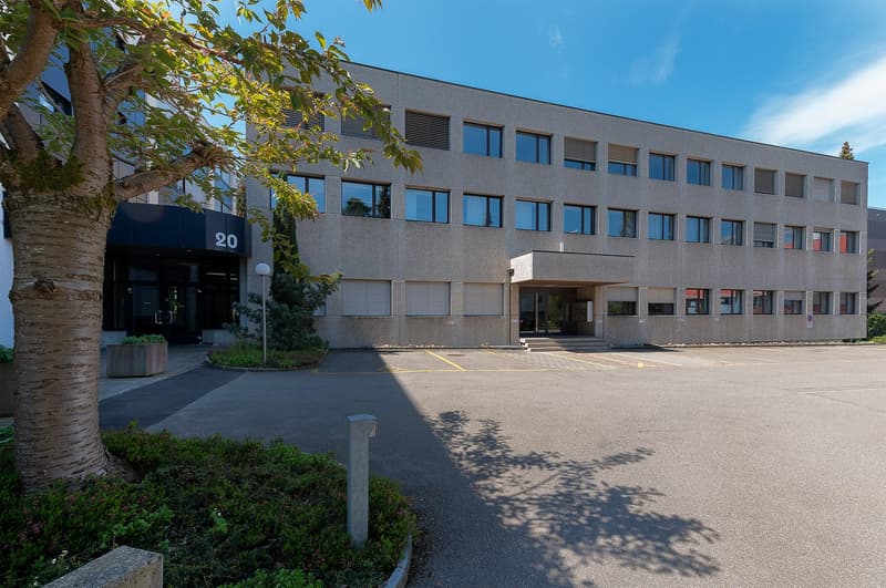 A multi-story office building with a gray exterior and large windows. The building has an entrance with a number '20' displayed above it. There is a paved parking area in front of the building, and some trees and landscaping around the entrance.