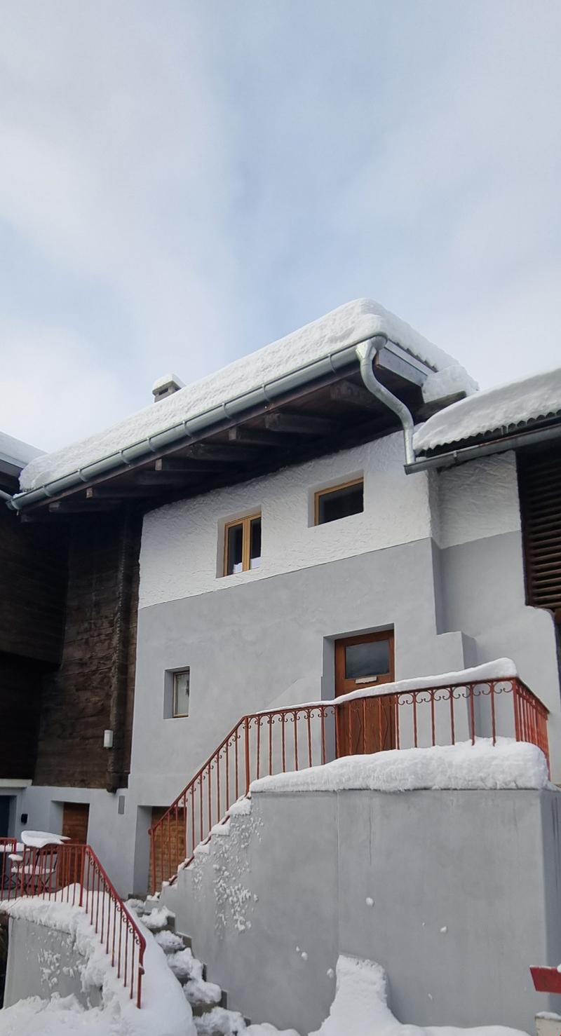 A two-story snow-covered house with white walls, a brown brick entrance, red railings, and multiple windows, with stairs leading up to the entrance. Snow-covered balconies are attached to the building.
