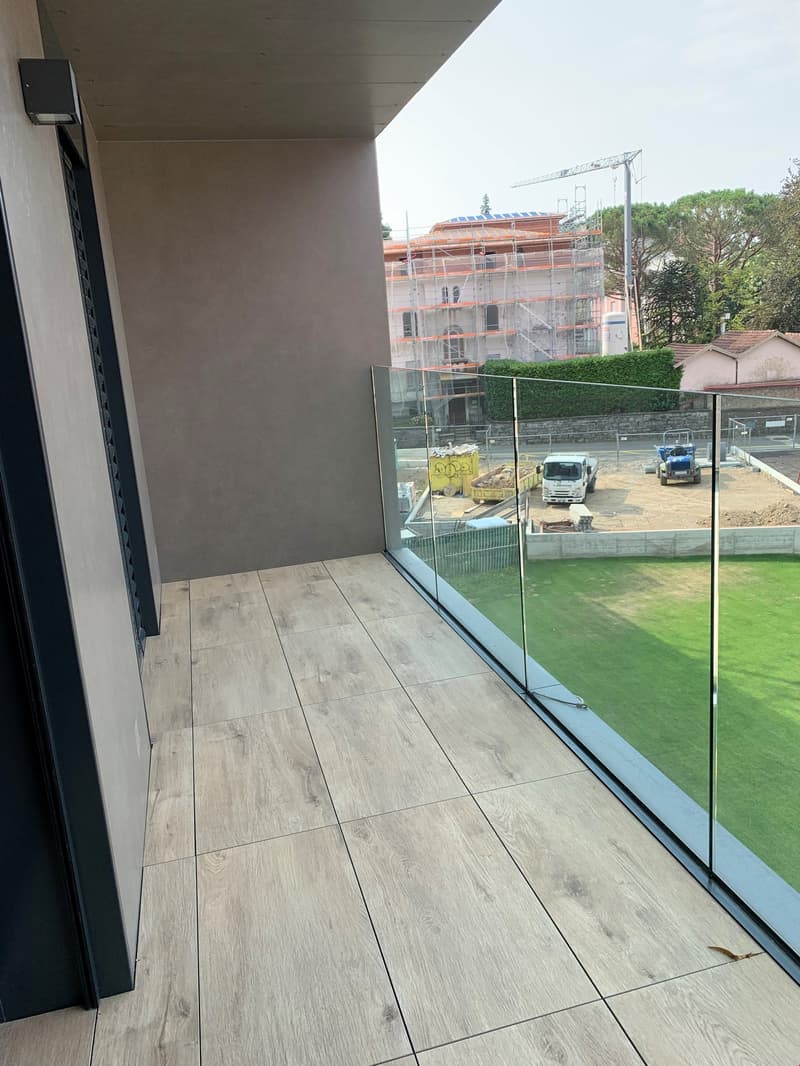 The image shows a balcony or terrace with a glass railing. The floor is covered in wood-like tiles, and there is a construction site visible through the glass railing.
