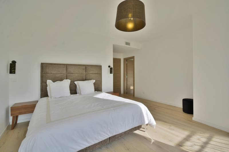 Bedroom with white walls, wooden floor, large bed with white sheets, two white pillows, two nightstands, a lamp, and a black ottoman.