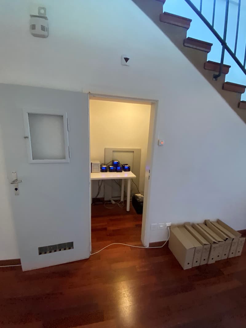 The image shows a room with a white door, a small window, and a table with some electronic devices on it. There are also some boxes or storage containers on the floor. The room has a hardwood floor and appears to be a utility or storage space.