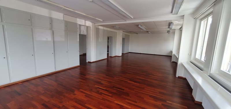 Large open office space with hardwood floors, white walls, and built-in storage cabinets along the walls. The room has high ceilings and large windows providing natural light.