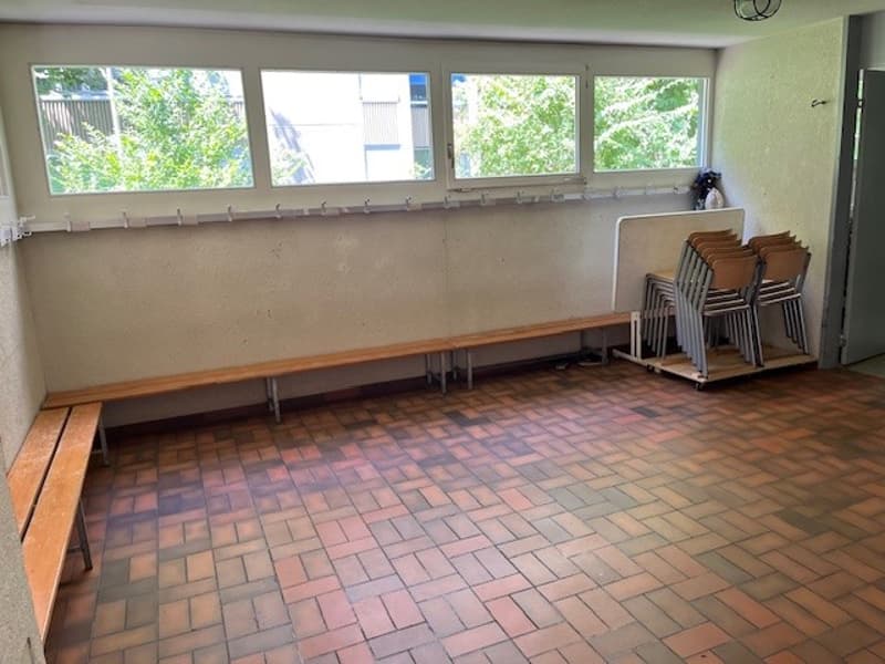 An empty room with red bricks flooring and two benches along the wall. Behind the benches is a wall with four large windows, revealing a view of trees outside. There is a stack of chairs in the corner near the door.