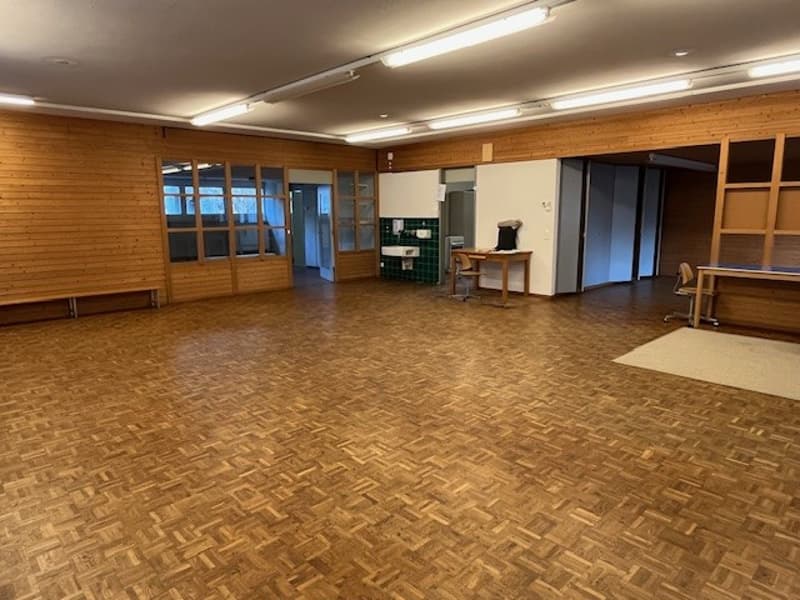 Empty wooden room with wood paneling, parquet floor, fluorescent lights, a desk, and a bench.