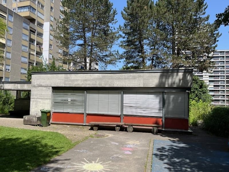 concrete and red walls building, raised ground floor, closed window shutters, small benches, green garbage can