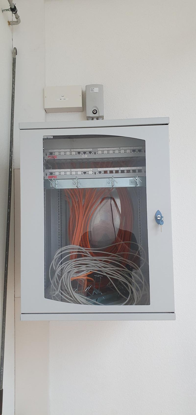 A white electrical box with a glass door, containing multiple cables and wires, mounted on a white wall.
