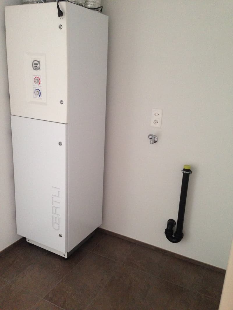 White appliance, likely a washing machine or dryer, with control panel visible. Tiled floor and plain white walls.