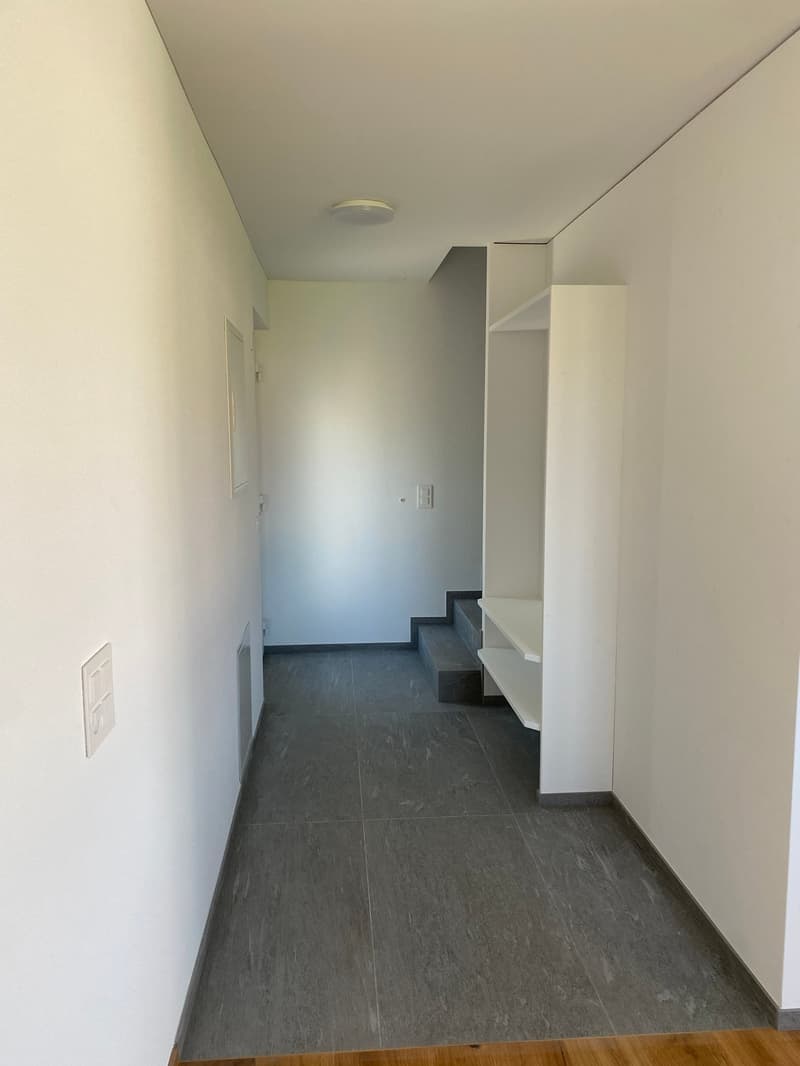 The image shows a hallway with white walls, a gray tile floor, and a set of stairs leading up. There is a light fixture on the ceiling and a door visible in the background.