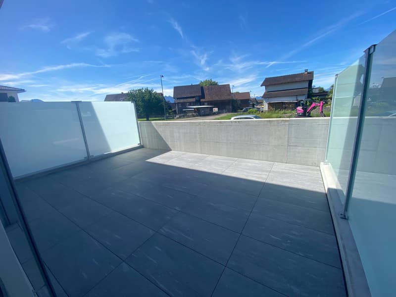 Concrete balcony with black tile flooring, overlooking a residential neighborhood with houses and a construction vehicle in the background. The sky is clear and blue with some wispy clouds.