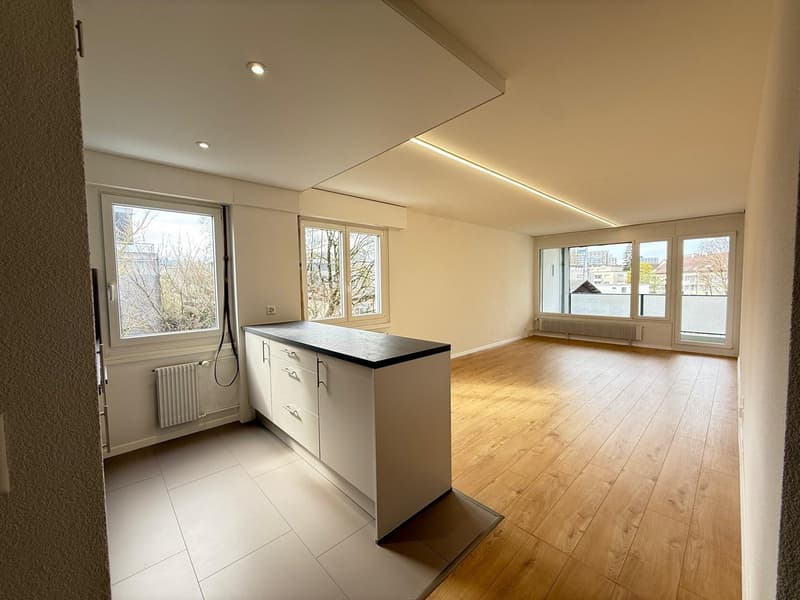 Empty kitchen with wooden floor, white cabinets, black countertop, windows, radiators, and ceiling lights