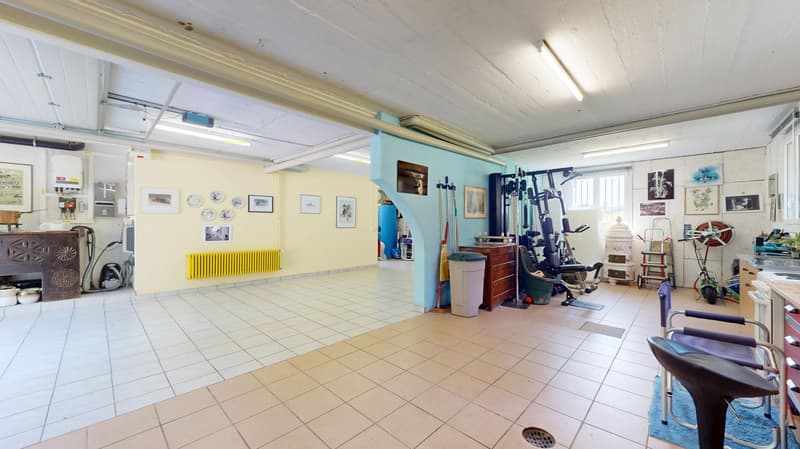 large open space, tiled flooring, yellow radiator, yellow wall, storage boxes, framed pictures, exercise equipment, trash can, blue chairs, brooms, lamps, wooden table, shelves with bowls, cabinets, electrical panels