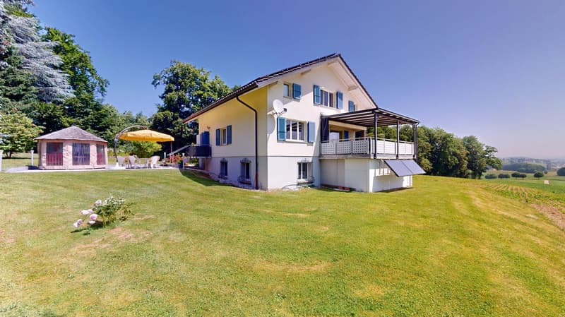 A two-story house with yellow walls, white shutters, a balcony, a solar panel, a garden with a yellow umbrella and a small gazebo