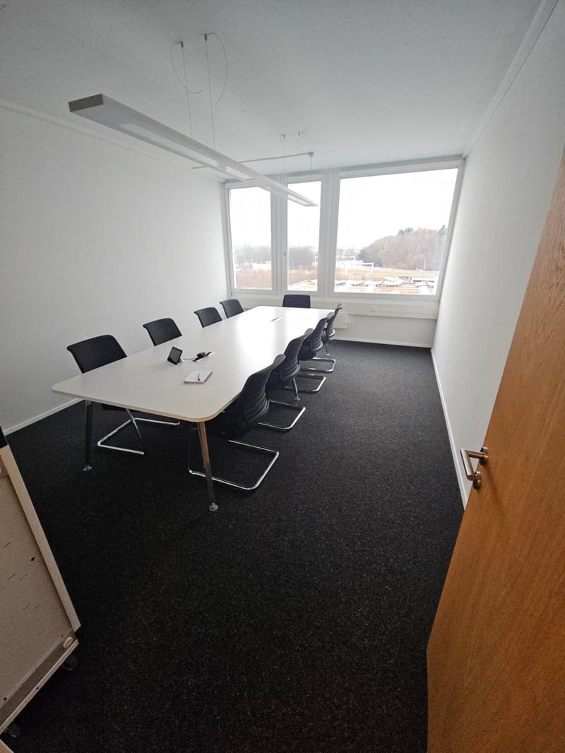 Long white table with several black office chairs, white walls, black floor, windows overlooking landscape, large windows