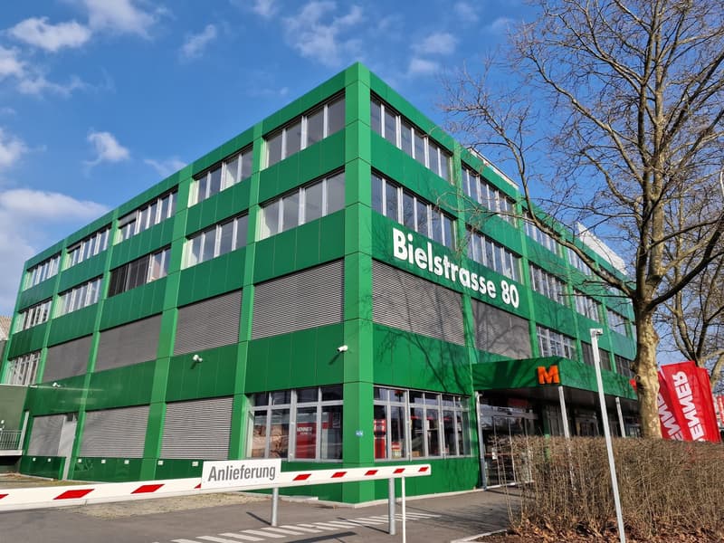 Office building with multiple floors, large windows, green paint, entry gate, building number '80', name 'Bielstrasse', street signage 'Anieferung', metal railings, surveillance cameras, bare tree, shrubs