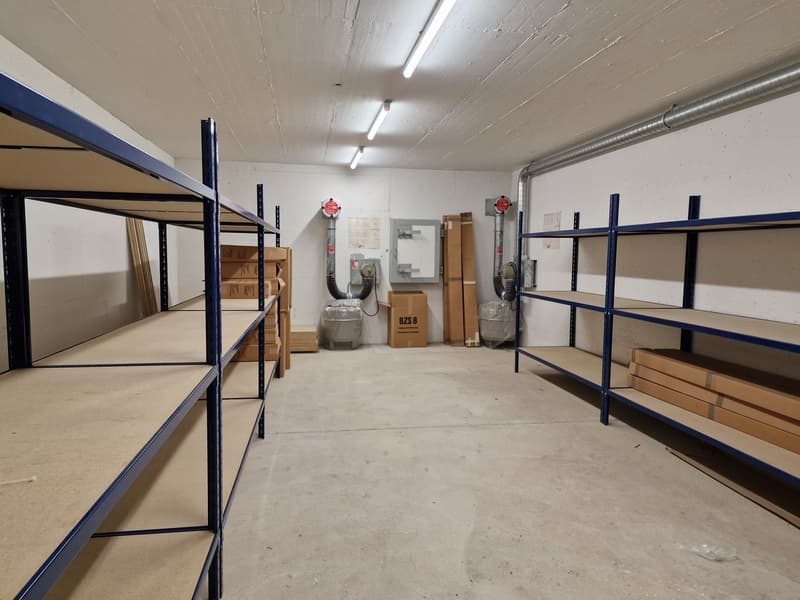 An empty room with white walls, metal shelves, and lighting, containing cardboard boxes and grey boxes