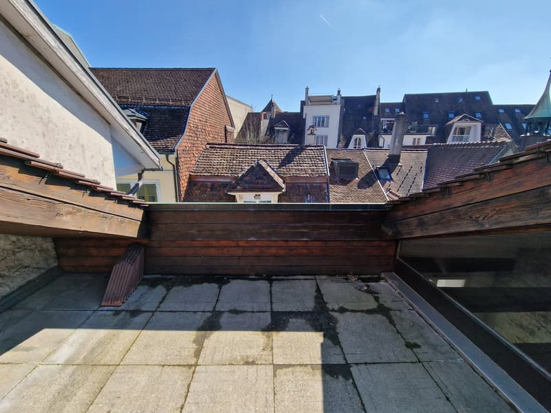 The image shows a view of a historic European city with traditional tile-roofed buildings. The buildings have a mix of architectural styles, with some having wooden beams and others having more modern facades. The rooftops are visible, with a mix of red t