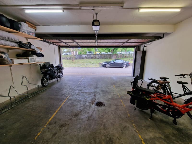 The image shows the interior of a garage. The garage has a concrete floor, shelves on the walls, and a motorcycle and other equipment stored inside. The garage door is open, revealing a car parked outside.