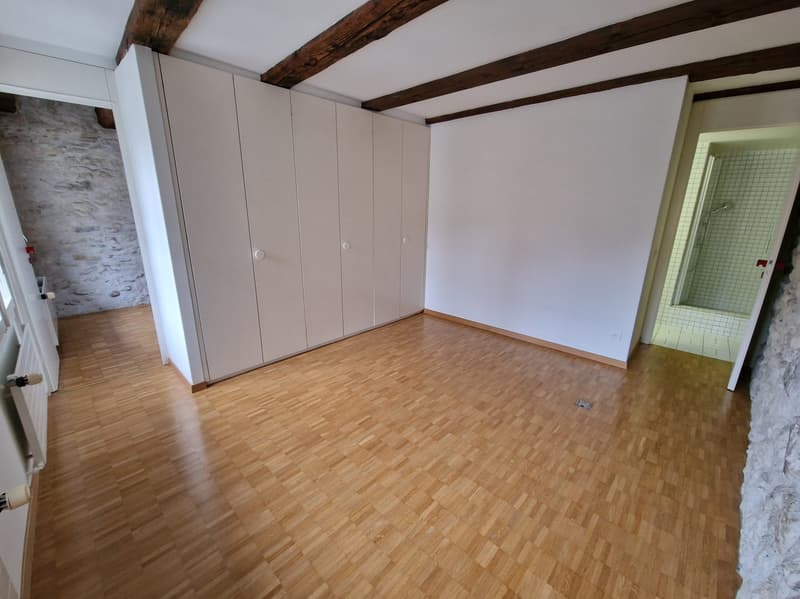 Spacious bedroom with hardwood flooring, white walls, and exposed wooden beams on the ceiling. The room features built-in wardrobes along one wall.