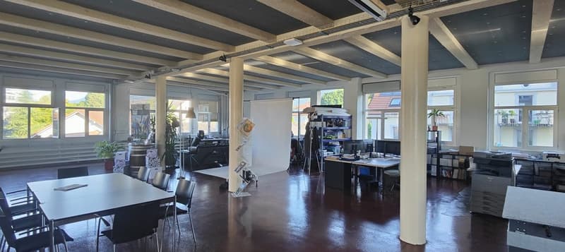 Large open-plan office space with high ceilings, exposed beams, and large windows. The space includes a conference table, desks, shelves, and various office equipment and supplies.