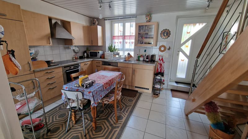 Wooden cabinets, stainless steel appliances, black stove, dining table with four chairs, white tiled floor, white walls, two lamps, window with blinds, countertop with sink, microwave, and various items.