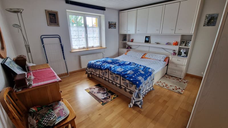 Bedroom with wooden floor, white walls, bed with blue comforter, bedside tables, radiator, window with curtains, cabinets, rug, framed picture on the wall, decorative items on shelves.