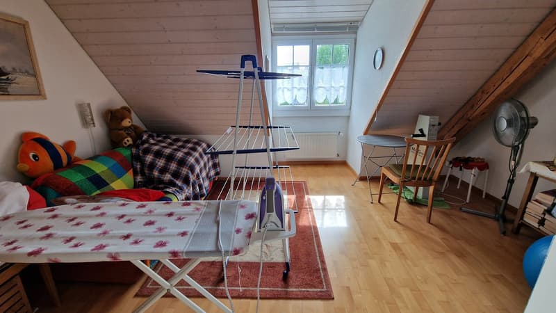 hardwood floors, white walls, small room, light from window, couch with blankets and toys, ironing table with iron, rug, desk, chair, small table, fan, books on small table