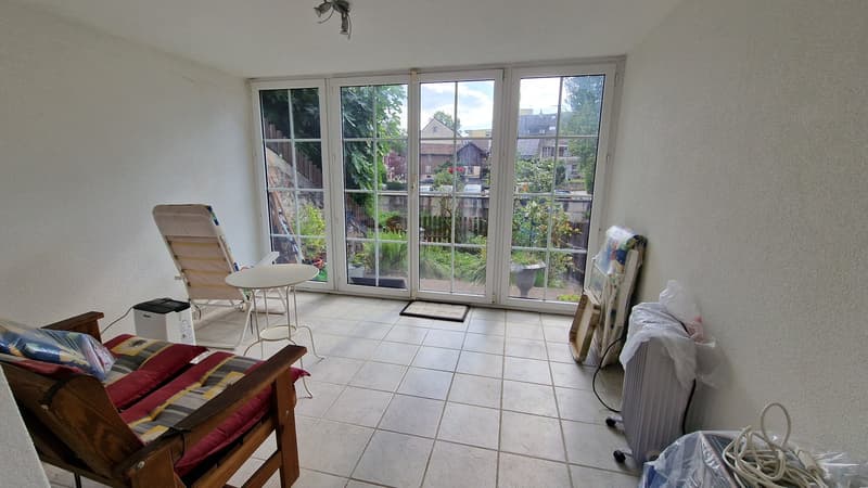 white walls, tile flooring, glass sliding doors leading to balcony, table, armchair, radiator, black power outlet, plastic bags, wooden furniture, hanging lamp