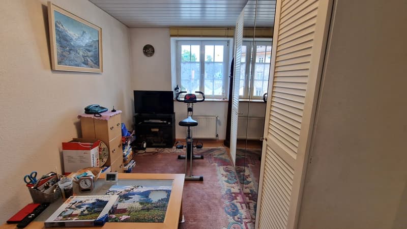 Room with beige walls, beige rug, beige desk, puzzle, clock, scissors, remote, books, file holder, basket, telephone, TV, exercise bike, storage boxes, shelves, radiator, picture frame, round wall decoration.