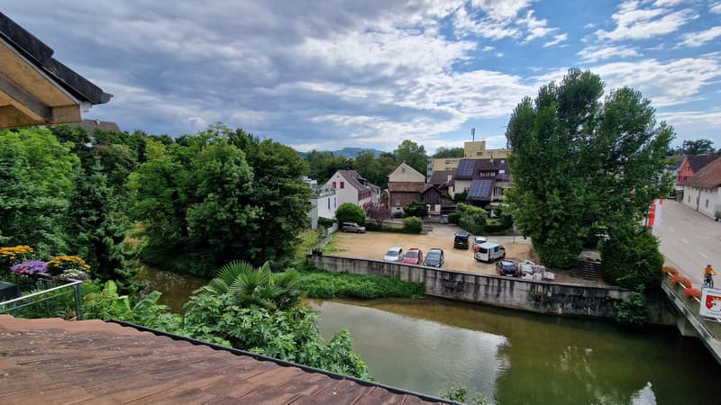 Houses with rooftops, greenery, river, parking area, road with traffic, cloudy sky