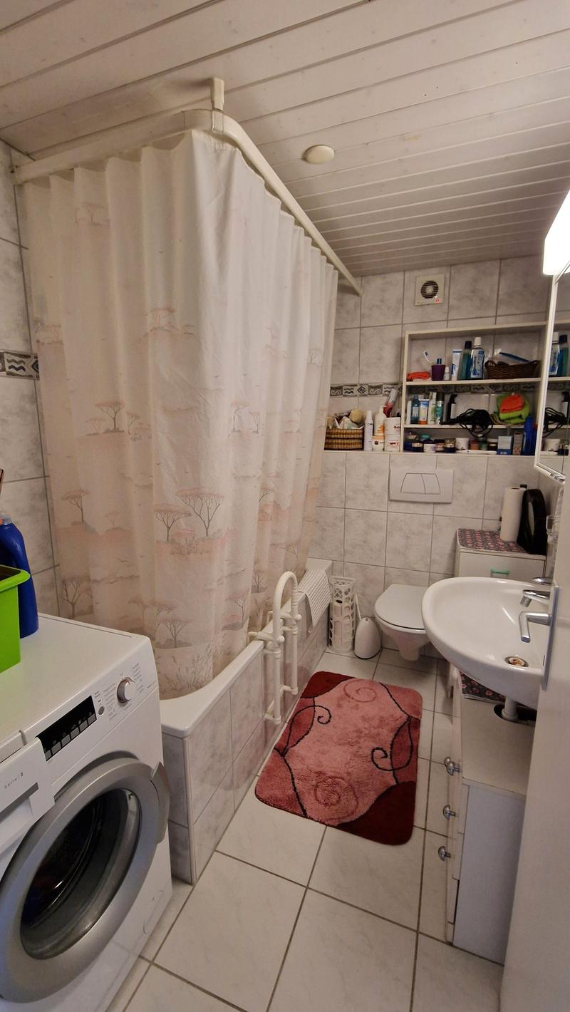 White tiled walls and floor, bath with shower curtain, white washing machine, white toilet, white sink, towel rack, shelves with toiletries and a basket, small pink rug on the floor, white radiator, green storage box on washing machine