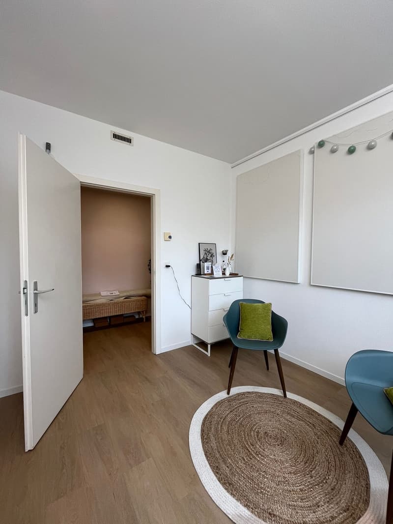 Empty room with wooden floor, white walls, open door, round rug, two chairs, white cabinet, and a small bed in the adjacent room.