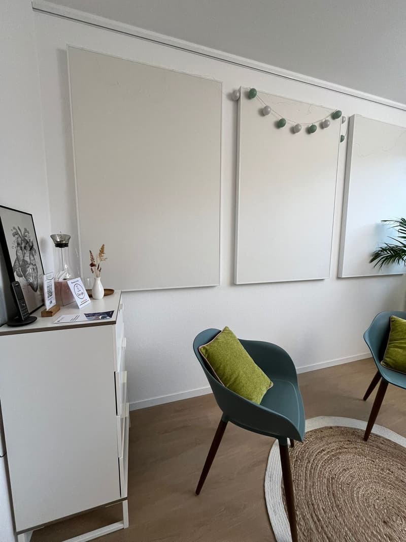 A room with a white wall, wooden floor, a white cabinet with a telephone, a vase, and a picture frame on top, two chairs with green pillows, and a round rug on the floor. There are three white boards mounted on the wall, and a string of green and white ba