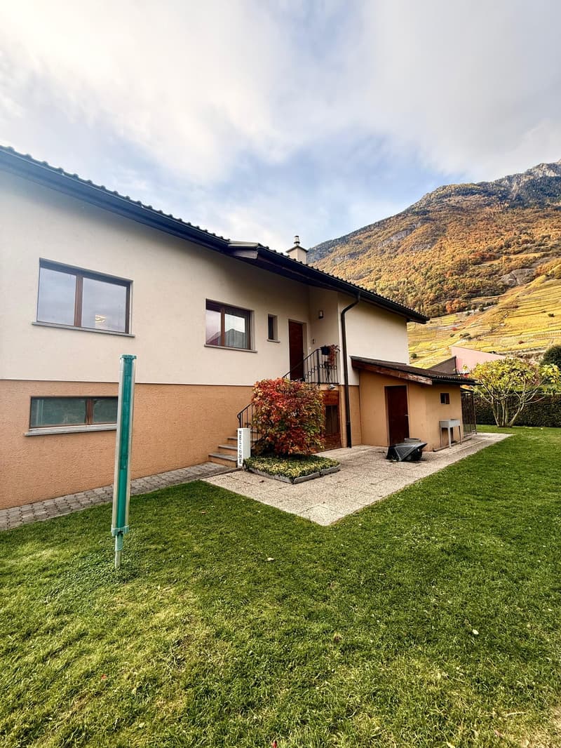 A house with 3 windows, white and beige walls, a chimney, a garden with plants, and a mountain in the background